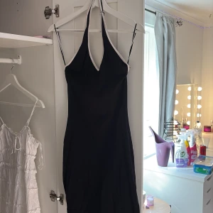 Svart halterneck klänning med vit kant - Snygg svart kortklänning med halterneck och smala axelband. Klänningen har en vit kant längs urringningen och är figurnära med en enkel, stilren design. Perfekt för dig som gillar minimalistisk och trendig stil.