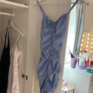 Ljusblå omlottklänning med knyt - Supersöt ljusblå kortklänning med smala axelband och knyt fram. Klänningen har en omlottliknande effekt och är sydd i ett krinklat bomullstyg som ger en avslappnad vibe. Perfekt för varma dagar och har en snygg rynkad detalj längs hela framsidan.  Liten fläck på rosetten pga hästen, borde gå bort i tvätt :)