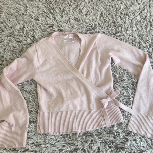 Ljusrosa omlottkofta från Gina Tricot - Superfin ljusrosa omlottkofta från Gina Tricot med knyt i sidan och ribbade muddar. Mjuk stickad känsla och långärmad modell som är perfekt för lager-på-lager. Snygg v-ringning och feminin rosettdetalj vid midjan.