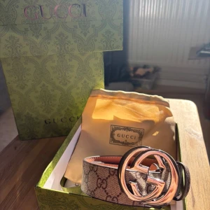 Beige bälte - Säljer ett bälte i beige och brunt med klassiskt monogrammönster och stor, rund metallspänne i silver. Bältet är gjort i skinn och har exklusiv känsla, perfekt för att lyfta vilken outfit som helst.