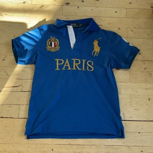 Blå Polo Ralph Lauren Paris pike - Blå pike från Polo Ralph Lauren med gul broderad logga och texten PARIS på bröstet. Snygga detaljer som broderat emblem och stor siffra 5 i vitt på ryggen och ärmen. Klassisk krage och korta ärmar.