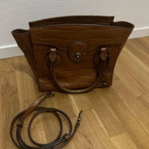 Brun handväska från Lauren Ralph Lauren - Snygg brun handväska i croco-mönstrat skinn från Lauren Ralph Lauren. Väskan har både handtag och avtagbar axelrem, samt ett stort fack med dragkedja. Perfekt för dig som vill ha en klassisk och trendig accessoar med lyxig känsla.