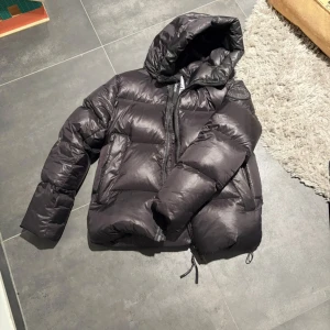 Svart dunjacka från Canada Goose - Säljer en svart pufferjacka från Canada Goose med huva och dragkedja. Jackan har klassisk logga på ärmen, stora fickor och justerbara snören nedtill. Perfekt för kalla dagar och riktigt snygg streetstil.       Är för närvarande i Tyskland men frakt möjlig✌️