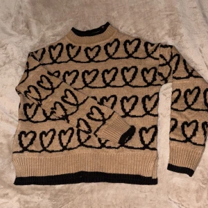 Beige stickad tröja med hjärtan - Mysig beige stickad tröja med svarta hjärtan över hela – ett unikt och charmigt plagg som sprider lite extra kärlek. Med svarta ribbade muddar vid hals, ärmslut och nederkant får den en stilren finish. Perfekt för dig som älskar bekväma plagg med en personlig touch ✨  ✔️ Från märket Sincerely Loved ✔️ Unikt hjärtmönster ✔️ Skön & varm – idealisk för både vardag och mys  💸 Jag säljer många fina plagg billigt – kika gärna på min sida för fler fynd! 🤝 Brukar också acceptera prisförslag.