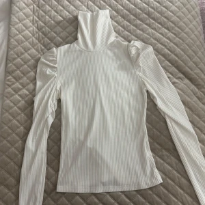 Vit ribbad polotröja med puffärm - Säljer en vit ribbad polotröja med långa ärmar och puff vid axlarna. Tröjan har en hög polokrage och är figurnära i modellen. Perfekt att styla med jeans eller kjol för en clean look.