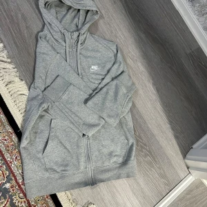 Grå hoodie från Nike med dragkedja - Snygg grå hoodie från Nike med huva och hel dragkedja framtill. Tröjan har två stora fickor och klassisk Nike-logga på bröstet. Perfekt för chill dagar eller när du vill ha en sportig look.