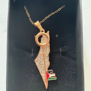 Palestina-halsband med glittrig karta - Unikt halsband med hängsmycke i form av Palestinas karta, täckt av gnistrande stenar och en nyckel. Finns i både guld- och silverfärg. Detaljer i rött, grönt, svart och vitt längst ner på hängsmycket. Perfekt statement-piece för dig som vill sticka ut.