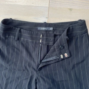 Svarta kostymbyxor med detaljer - Svarta kostymbyxor med smala vita ränder. Jag har använt dessa byxor en gång men köpt second hand så därför kan ej svara helt på hur använda de är. Inga defekter syns. De är från märket InWear Essentails och storlek 34. De sitter löst nere och lågmidjat. Byxorna är i en blandning av kostymbyxor matrial och stretch/tights aktigt skulle jag säga. 