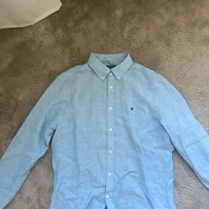 Ljusblå linneskjorta från Morris - Snygg ljusblå skjorta från Morris i 100% linne. Klassisk passform med button down-krage, vita knappar och broderad logga på bröstet. Perfekt för dig som gillar stilrena och fräscha plagg med en avslappnad vibe.