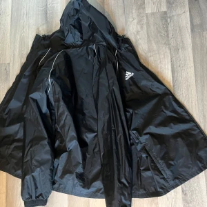 Svart vindjacka från Adidas XL - Svart vindjacka från Adidas i storlek XL. Jackan har huva, vita detaljer och Adidas-logga på bröstet. Tillverkad i lätt polyester med elastiska muddar vid ärmsluten. Perfekt för blåsiga dagar och sportiga outfits.