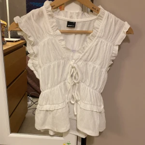 Vit volangtopp med knyt från Gina Tricot - Supersöt vit topp från Gina Tricot i storlek XS. Toppen har volangdetaljer, v-ringning och knyt framtill. Ärmlös modell med lätt struktur i tyget som ger en luftig känsla. Perfekt för varma dagar när du vill känna dig extra fin.
