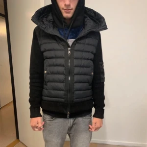 Svart Moncler pufferjacka med stickade ärmar - Säljer en svart pufferjacka från Moncler med stickade ärmar och huva. Jackan har dragkedja framtill, fickor med tryckknappar och en extra ficka med dragkedja på ärmen. Klassisk Moncler-logga på vänster ärm. Perfekt för kyliga dagar. 