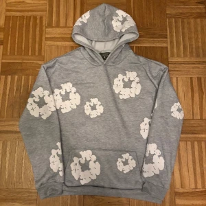 Grå hoodie från Denim Tears med blommor - Grå hoodie från Denim Tears med vita blommiga tryck över hela tröjan. Klassisk huva och känguruficka framtill. Mjuk insida och relaxed fit, perfekt för chill dagar. Snygg och unik design som sticker ut.