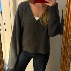 Mörkgrå kofta med v-ringning - Mysig mörkgrå kofta med v-ringad hals och knappar framtill. Koftan har långa, lite vidare ärmar och ribbade muddar vid ärmslut och nederkant. Perfekt för lager på lager och enkel att styla till jeans eller kjol.