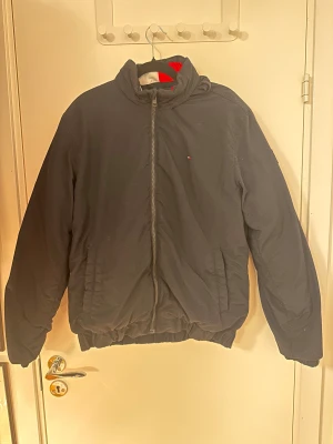 Svart pufferjacka från Tommy Hilfiger - Svart pufferjacka från Tommy Hilfiger med diskret logga på bröstet. Jackan har hög krage, två sidofickor och hel dragkedja framtill. Den har också en luva som är inbyggd i kragen. Materialet är polyamid på utsidan och polyesterfoder. Perfekt för kalla dagar och har en clean, enkel look.