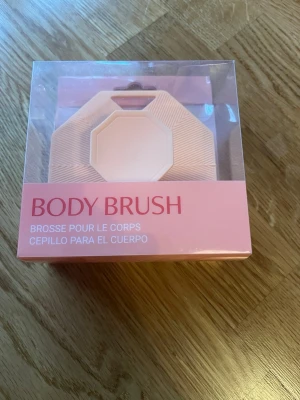 Rosa body brush i silikon - Säljer en rosa body brush i silikon med geometrisk form och praktiskt handtag. Perfekt för att applicera och arbeta in hudvårdsprodukter eller exfoliera kroppen. Kommer i snygg förpackning med modern design.