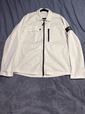 Beige overshirt från Stone Island - Snygg beige overshirt från Stone Island med klassisk patch på ärmen. Jackan har rak passform, dragkedja framtill, bröstficka med knapp och en vertikal ficka med dragkedja. Tillverkad i bomull och perfekt för lager-på-lager. Stilren och enkel att matcha. Har knappast använts har bara hängt i garderoben. Köpte den på NK i Stockholm. Ny priset på jackan är 3,700kr när jag köpte den i butik. Har kvitto bevis på jackan.