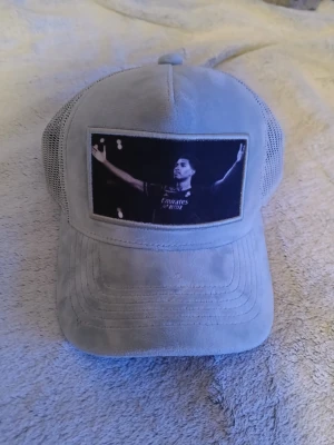 Grå bellingham mockakeps med mesh och tryck - Snygg grå keps i mockaimitation med mesh på sidorna och baksidan för extra ventilation. Framtill finns ett svart tryck med motiv av en person med utsträckta armar. Justerbar passform med snapback. Perfekt för dig som vill ha en unik och cool accessoar.