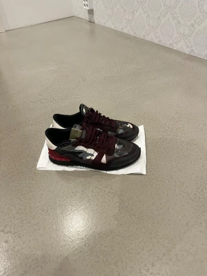 Valentino Rockrunner sneakers camouflage - Säljer ett par Valentino Rockrunner sneakers med vinröda och svarta detaljer, samt coolt camouflage-mönster i grått, vitt och brunt. Skorna har snörning, platt sula och rund tå. Materialmix med mocka och textil. Perfekt för dig som gillar statement pieces. Hör av med fler frågor. 