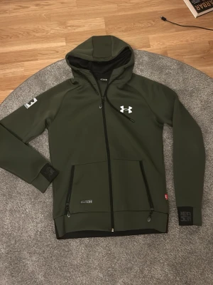 Under Armour Hoodie - olivgrön - Fet under armour zip med reflexer. Den är helt oanvänd, storlek M på tagsen men sitter som en S. Pris kan diskuteras vid snabb affär.