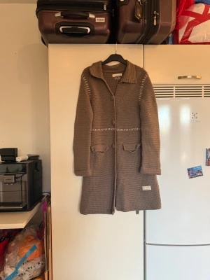 Beige lång kofta från Odd Molly - Säljer en brun lång kofta från Odd Molly med grovstickad struktur och dekorativa vita detaljer vid axlar och midja. Koftan har stora knappar framtill, krage och två fickor med lock. Perfekt för lager-på-lager under kyliga dagar. Storlek 1 som enligt deras storleksguide motsvarar en S. Mer använd men fortfarande fin. Kan dock ha tecken på användning. 