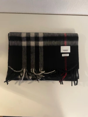 Rutig svart Burberry kashmirväst - Snygg väst från Burberry i klassiskt rutigt mönster med svarta, gråa och röda toner. Västen är tillverkad i 100% kashmir och har fransar längs nederkanten. Märket och materialetiketten syns tydligt på framsidan. Perfekt för dig som gillar lyxiga och tidlösa plagg.