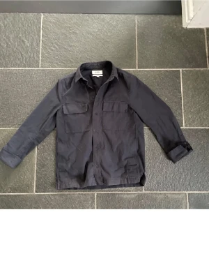 Marinblå Overshirt från COS - Snygg marinblå overshirt från COS med långa ärmar och knappar framtill. Perfekt för lager-på-lager stil. Två praktiska bröstfickor ger en cool touch. Passar både till vardags och mer uppklädda tillfällen. Inga hål eller defekter, vid frågor eller funderingar hjälper jag gärna till. Mvh Olof.