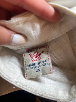 True Religion vita jeans med paljettdetalj - Säljer ett par vita jeans från True Religion med ikonisk logga i silverpaljetter på bakfickan och snygg knappdetalj. Jeansen är tillverkade i USA av bomullstyg och har en klassisk passform med låg midja. Perfekt för dig som gillar statement-plagg och coola detaljer.