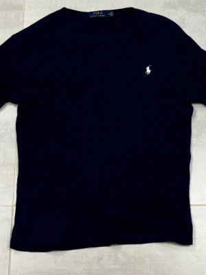 Mörkblå stickad tröja Polo Ralph Lauren - Snygg mörkblå stickad tröja från Polo Ralph Lauren storlek L med klassisk rund hals och vit broderad logga på bröstet. Tröjan har långärmade ribbade muddar och är gjord i ett mjukt stickat material som passar perfekt till höst och vinter.