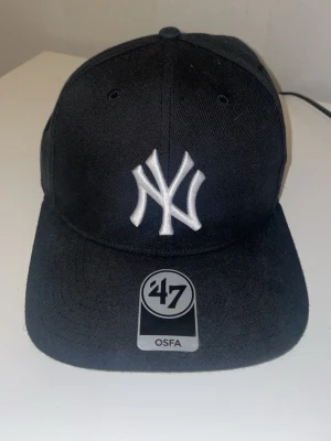 Svart New York Yankees keps från '47 - Säljer en svart New York Yankees keps från märket '47 med klassisk böjd skärm och broderad vit NY-logga framtill. Kepsen är tillverkad i en blandning av ull och syntet, vilket ger en mjuk och slitstark känsla. Perfekt för dig som vill ha en sportig och stilren look.