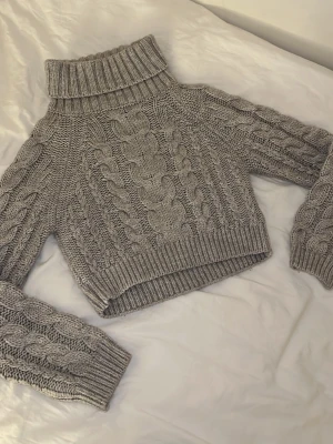 Grå stickad polotröja med kabelmönster - Säljer en grå stickad polotröja med grovt kabelmönster och ribbade muddar. Tröjan har hög polokrage och långärmade ärmar, perfekt för kyliga dagar. Den är lite croppad i modellen och har en chunky vibe som är både snygg och bekväm.