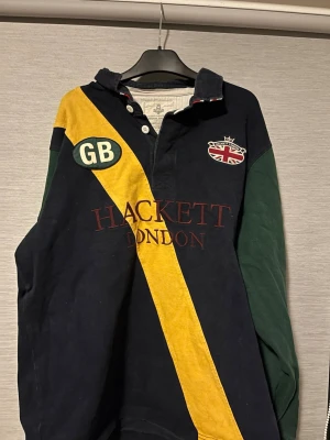 Hackett London rugbytröja XL som passar M - Snygg rugbyinspirerad långärmad skjorta från Hackett London i storlek XL men som var krympt till M. Marinblå bas med gröna ärmar, gul diagonal rand och broderade detaljer som GB-märke och brittisk flagga. Kraftig bomullskvalitet och klassisk krage med knappar framtill. Hör av dig vid intresse!🤩🤩