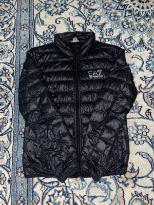 Svart pufferjacka från EA7 Emporio Armani - Säljer en svart pufferjacka från EA7 Emporio Armani med stilren logga på bröstet. Jackan har quiltad design, dragkedja framtill och långärmade ärmar. Materialet är glansig polyester som ger en modern vibe. Perfekt för dig som vill ha en snygg och lätt jacka med streetkänsla.