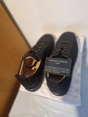 Svarta sneakers från Christian Louboutin - Säljer ett par svarta sneakers från Christian Louboutin med snyggt broderad logga på hälen och ikonisk röd sula. Skorna har snörning, rund tå och är tillverkade i mjuk mocka med skinnfoder. Perfekt för dig som vill ha något exklusivt och stilrent.