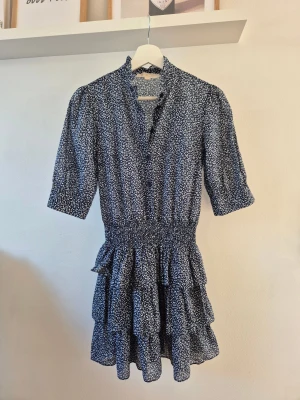 Klänning Michael Kors | Nypris 1 700kr - Supersöt kortklänning från Michael Kors i marinblått med små vita blommor. Klänningen har volanger, smock i midjan, knappar framtill och trekvartsärm. Tillverkad i mjuk polyester som ger ett luftigt och flowy intryck. Perfekt för dig som gillar romantiska detaljer och blommigt.