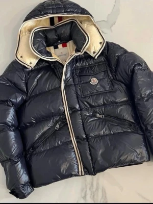 Svart Moncler pufferjacka med huva - Snygg svart pufferjacka från Moncler med glansig finish och avtagbar huva. Jackan har en stor bröstficka med Moncler-logga, två snedställda fickor med dragkedja och krämfärgat foder med röd, vit och marinblå rand. Perfekt för kalla dagar. MÖTS BARA ‼️