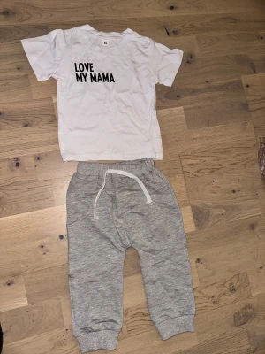 Grå mjukisbyxor med snörning Set med T-shirt och Byxor - Säljer ett par grå mjukisbyxor med vit snörning i midjan och ribbade muddar vid bensluten. Byxorna har en avslappnad passform och är gjorda i mjukt bomullsmaterial, perfekta för chill dagar. Passar dig som gillar enkel och bekväm stil.Ett sött set bestående av en vit t-shirt med texten "LOVE MY MAMA" och ett par grå byxor med dragsko. 1år