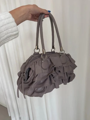 Lila handväska med volanger - Säljer en lila handväska med snygga volangdetaljer och flätade handtag i matchande färg. Väskan har dragkedja upptill och ett rymligt fack inuti. Materialet är mjukt skinnliknande syntet och detaljer i silver. Perfekt storlek för det viktigaste. Lite av färgen har lossnat runt väskan, samt ena handtaget sitter lite löst (kolla bild)!