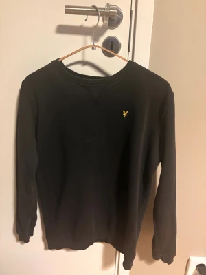 Svart sweatshirt från Lyle & Scott - Svart sweatshirt från Lyle & Scott Junior med klassisk rund hals och gul broderad logga på bröstet. Tröjan har ribbade muddar vid ärmslut och nederkant, samt långärmad passform. Perfekt för dig som gillar stilrena och enkla plagg.