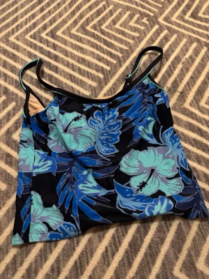 Blommig blå linnetopp med smala band - Snygg linnetopp med tropiskt blommönster i blå och turkosa nyanser på svart botten. Toppen har smala axelband och är croppad i modellen. Perfekt för varma dagar och ger en fräsch vibe till din outfit. Linnet är gjort i baddräkts material. Märke eller storlek står inte men passar mig som är S.