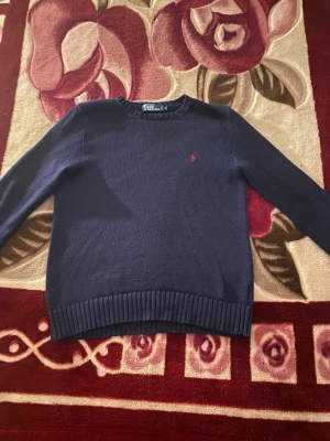 Mörkblå stickad tröja Polo Ralph Lauren - Snygg mörkblå stickad tröja från Polo Ralph Lauren i storlek S. Tröjan är gjord av 100% bomull och har ett klassiskt rakt snitt med ribbade muddar och nederkant. På bröstet finns den ikoniska röda Polo-loggan broderad. Perfekt för dig som gillar stilrena och tidlösa plagg.