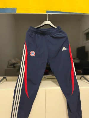 Adidas Bayern München träningsbyxor - Säljer ett par mörkblå Adidas Bayern München träningsbyxor med vita och röda detaljer längs sidorna. Byxorna har elastisk midja och klubbmärke på ena benet samt Adidas-logga på andra. Perfekta för fotboll eller chill.