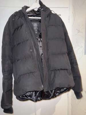 Canada Goose Macmillan Parka - Svart pufferjacka från Canada Goose med huva och broderad logga på ärmen. Dustbag medföljer. Jackan har dragkedja, knappar och snörning vid huvan. Fodrad insida och två sidofickor. Perfekt för kalla dagar och har en clean, stilren look. 