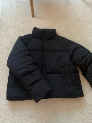 Svart pufferjacka från Monki - Svart pufferjacka från Monki med hög krage och grov dragkedja framtill. Jackan har en boxig form och är vadderad för extra värme. Perfekt för kalla dagar och har långa ärmar samt sidofickor. Enkel och stilren design som funkar till många outfits.