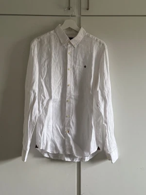 Vit linneskjorta från Morris Stockholm - Säljer en stilren vit skjorta från Morris Stockholm i ren linne. Skjortan har klassisk krage, knappar framtill och ett broderat emblem på bröstet. Perfekt för dig som gillar en clean och fräsch look. Passar dig som vill ha något luftigt och snyggt. 100% linne