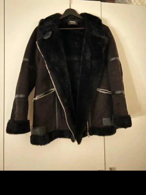 Svart pälsjacka med dragkedjor - Snygg svart pälsjacka från Ricano med fluffig krage och muddar. Jackan har två snedställda dragkedjor fram och detaljer i skinn på ärmar och nederkant. Perfekt för dig som gillar en cool och varm look under höst och vinter.