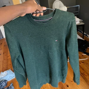 Mörkgrön stickad tröja Tommy Hilfiger - Snygg mörkgrön stickad tröja från Tommy Hilfiger med diskret logga broderad på bröstet. Tröjan har rund halsringning och långa ärmar, perfekt för dig som gillar klassisk stil med en modern touch. Passar dig som vill ha en enkel men stilren look.