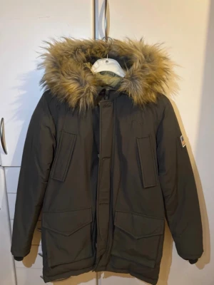 Mörkgrön parkajacka från Svea med fuskpäls - Parkajacka från Svea i mörkgrönt med stor huva och fluffig fuskpäls. Jackan har flera praktiska fickor, broderad Svea-logga på bröstet och ett patch med amerikansk flagga på ärmen. Insidan är ljusbeige och jackan stängs med dragkedja och knappar.