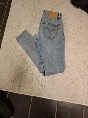 Ljusblå jeans från Tiger of Sweden - Säljer ett par ljusblå jeans från Tiger of Sweden med klassisk femficksdesign och snygga kontrastsömmar. Jeansen har en slim passform och är tillverkade i mjukt denimtyg som sitter skönt hela dagen. Perfekt för dig som gillar stilrena och moderna jeans.