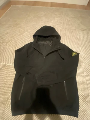 Svart vindjacka Stone Island XS - Svart vindjacka från Stone Island med huva och dragkedja framtill. Jackan har två sidofickor med dragkedja och klassisk Stone Island patch på vänster ärm. Tillverkad i Italien och gjord i polyester, perfekt för dig som gillar streetwear.